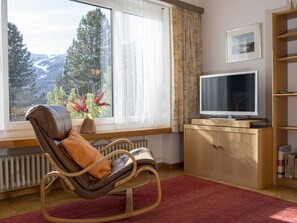Une pièce avec un fauteuil à bascule, un téléviseur, une bibliothèque et une fenêtre donnant sur des arbres et des montagnes.