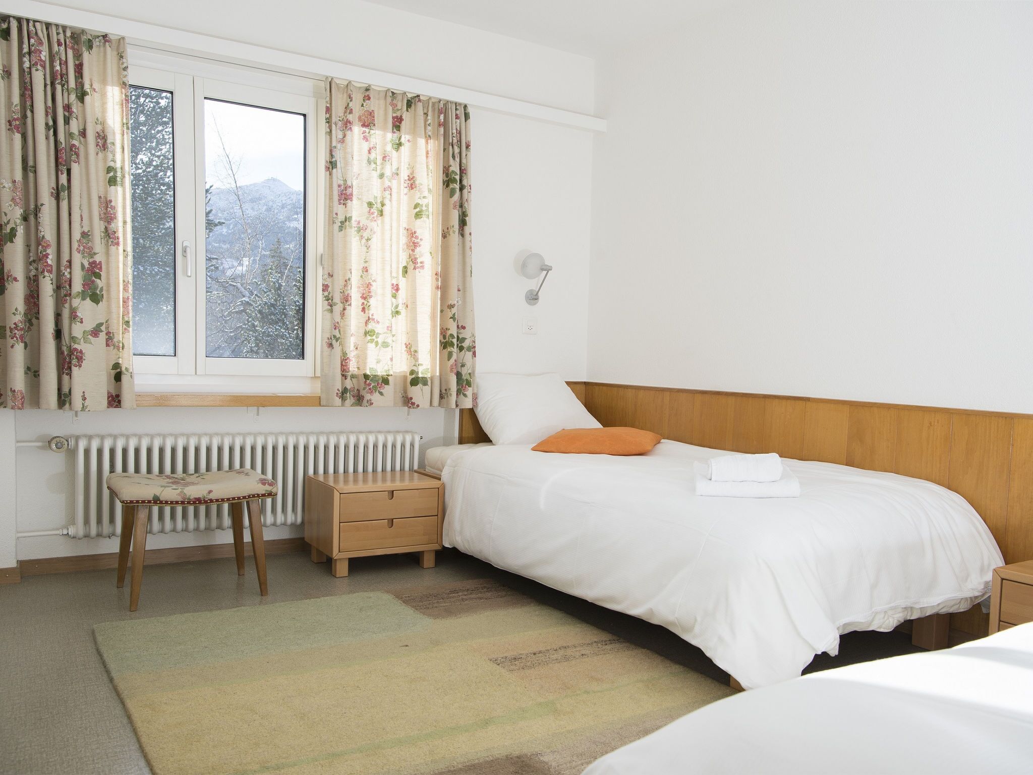Un dormitorio con cama, mesita de noche, silla, radiador y ventana con cortinas.