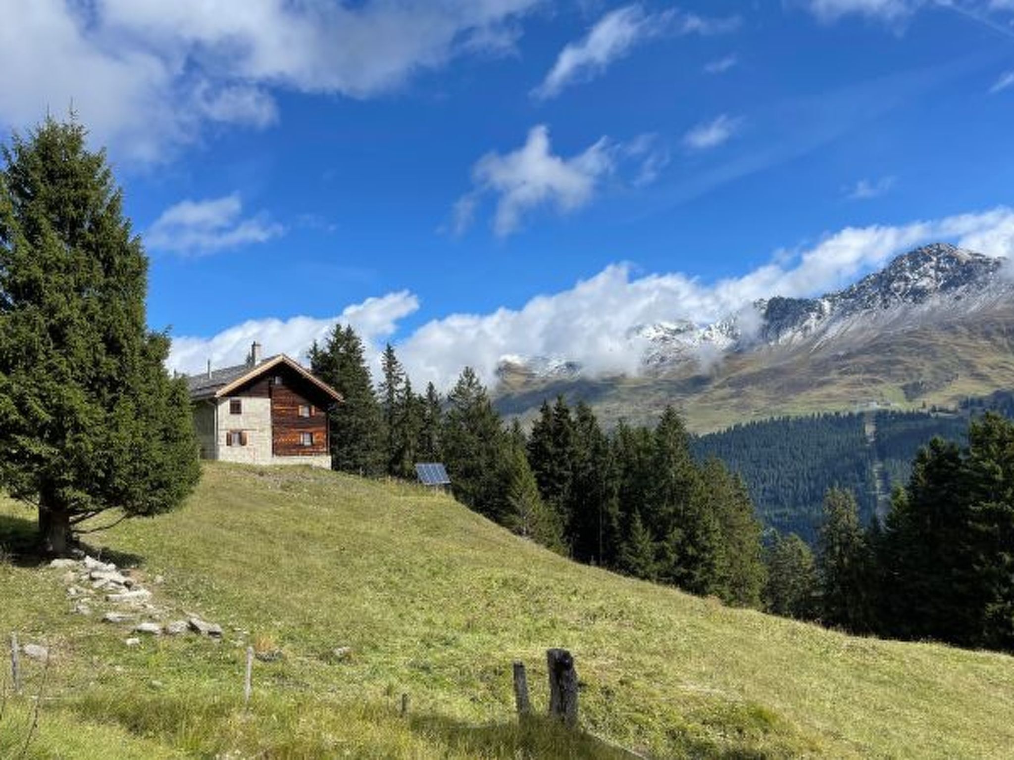 A wooden cabin with a sloped roof, surrounded by a grassy hill, trees, and a mountain range in the background.