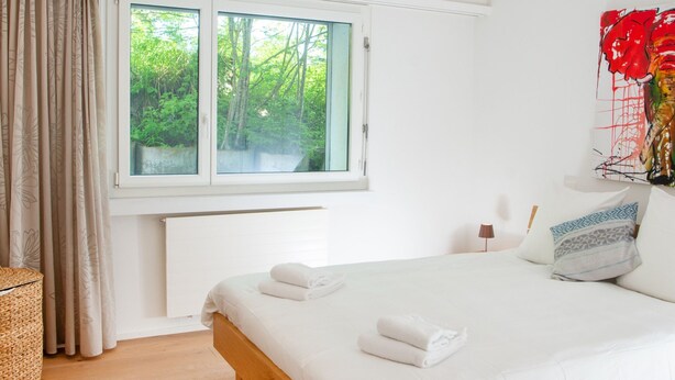 Quarto com uma janela ampla, cama com roupa de cama branca e uma obra de arte na parede.