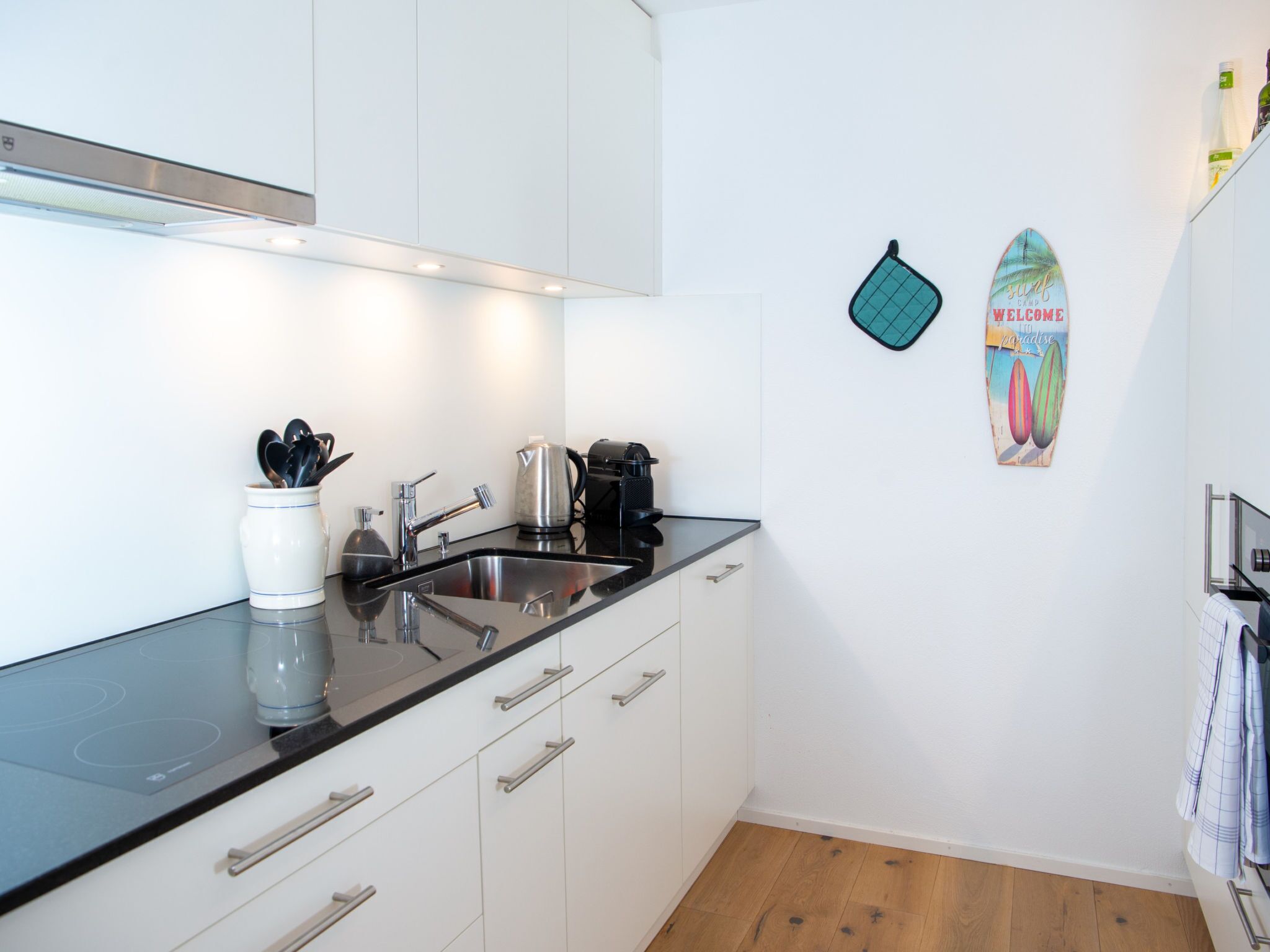 Una cocina moderna con armarios blancos, encimera negra y pileta de acero inoxidable. Hay una tetera, una cafetera y un tostador sobre la encimera.