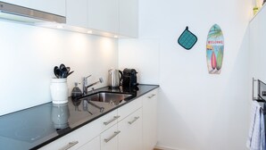Uma cozinha moderna com armários brancos, bancada preta e uma pia de aço inoxidável. Há uma chaleira, uma cafeteira e uma torradeira na bancada.