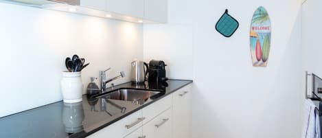 Une cuisine moderne avec des meubles blancs, un plan de travail noir et un évier en acier inoxydable. On y trouve une bouilloire, une machine à café et un grille-pain sur le plan de travail.