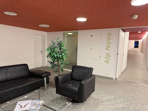 A waiting area with two black leather chairs, a glass coffee table with magazines, a potted plant, and a wall with the text 'Alp Nova'. - 1614 La Riva 401 by Interhome (Lenzerheide)