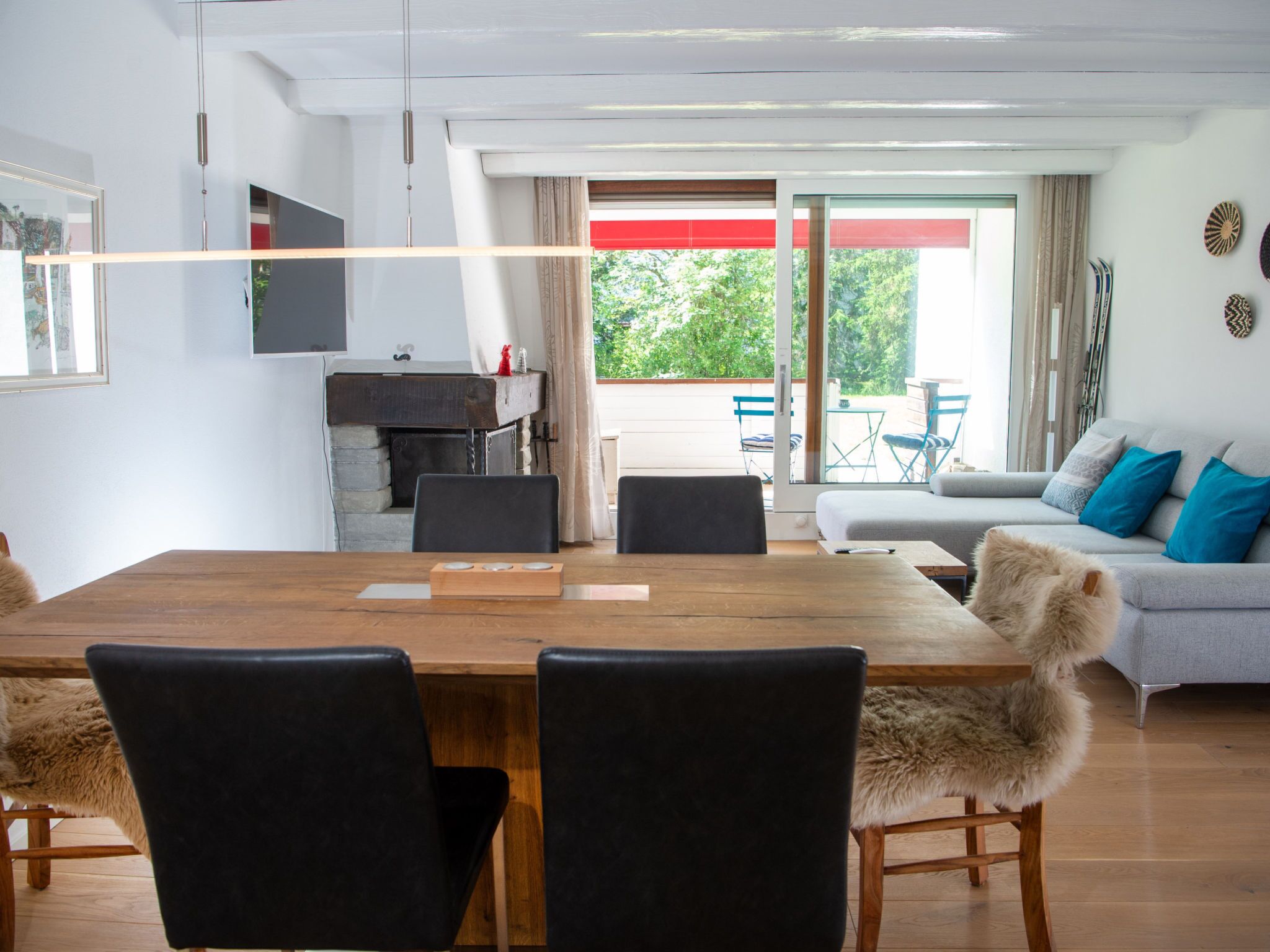 A modern living room with a wooden dining table, black chairs, a grey sofa with blue cushions, and a view of a balcony with outdoor furniture.