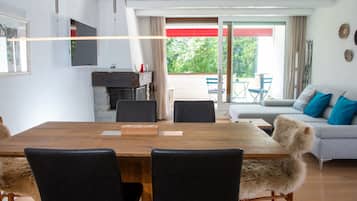 A modern living room with a wooden dining table, black chairs, a grey sofa with blue cushions, and a view of a balcony with outdoor furniture.