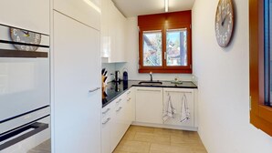 Eine kleine Küche mit weißen Schränken, einem eingebauten Backofen, einem Fenster mit Aussicht und einer Wanduhr.