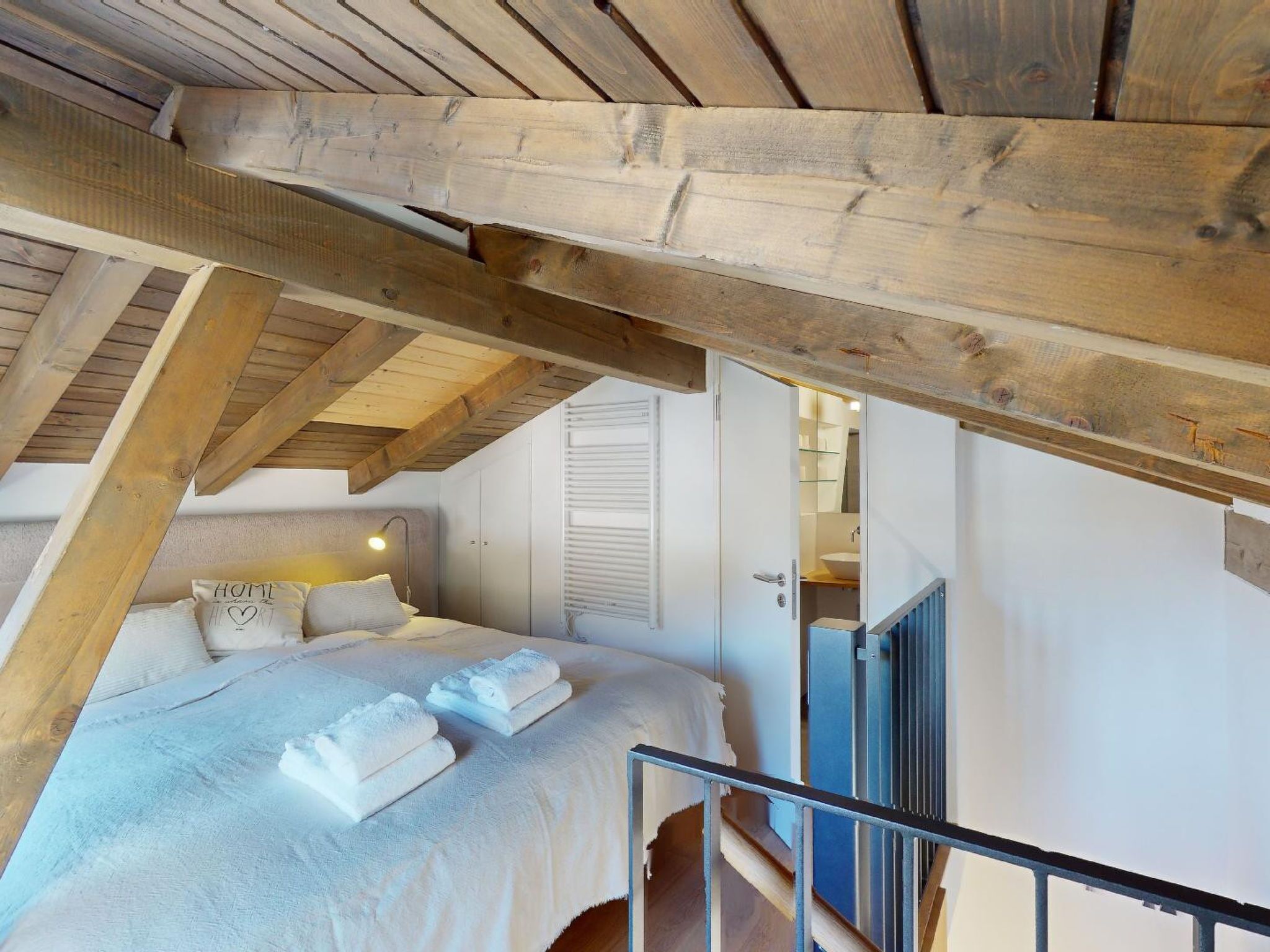 Une chambre avec un plafond en pente, un lit recouvert de draps blancs, une lampe de chevet et une vue sur la salle de bain par une porte ouverte.