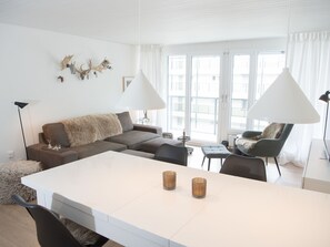 Ein modernes Wohnzimmer mit einer Couch, einem Esstisch und zwei Stühlen. Auf dem Tisch stehen zwei Lampen und an der Wand ein Kronleuchter.