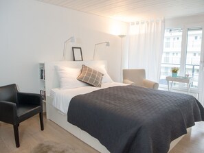 Ein modernes Schlafzimmer mit einem großen Bett, einem Nachttisch, einem Sessel und einem Fenster mit Vorhängen.