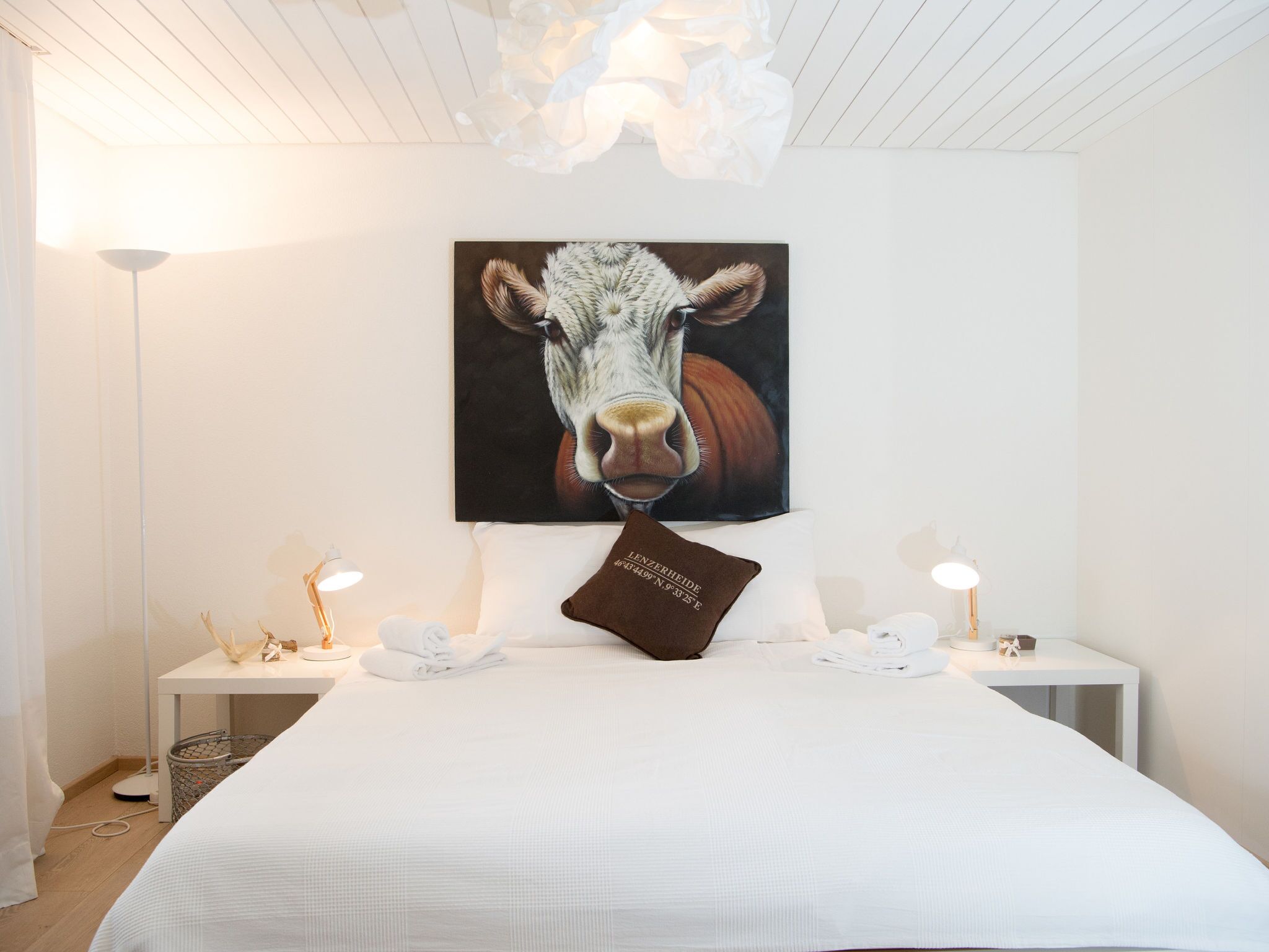 A hotel room with a large bed, a painting of a cow on the wall, a pillow with text, and bedside lamps.