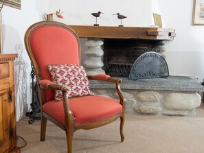 A wooden armchair with a red cushion and a floral pillow, positioned in front of a stone fireplace with a metal screen. - 1615 Bot La Pala by Interhome (Lenzerheide)