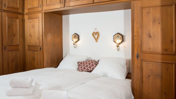 Een tweepersoonsbed met witte lakens en twee kussens, een hartvormige wanddecoratie en twee wandlampen.