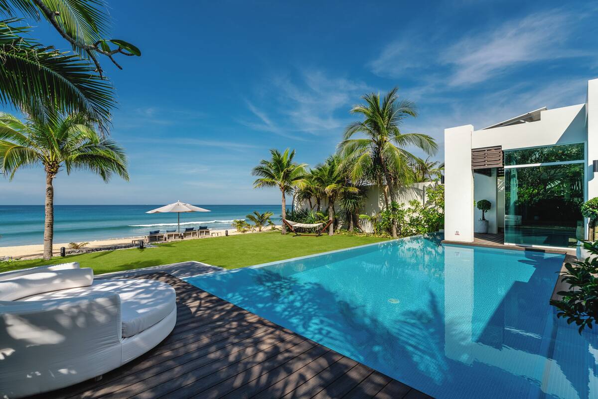 A modern pool area with a view of the ocean, palm trees, and a white building with large windows.