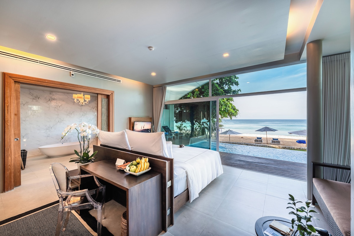 A modern hotel room with a large bed, a desk with a fruit bowl, a view of the beach, and a glass-enclosed balcony.