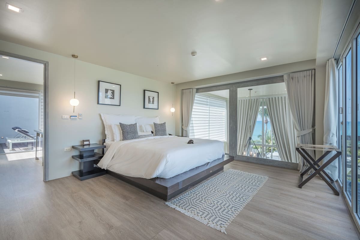A modern bedroom with a large bed, a built-in bench, a small table, and a view of the outdoors through large windows.