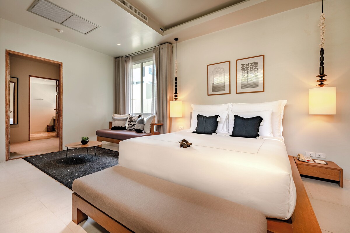 Quarto de hotel moderno com uma cama grande, mesas de cabeceira, um banco, uma pequena mesa com uma planta e vista para o exterior através de uma janela ampla.