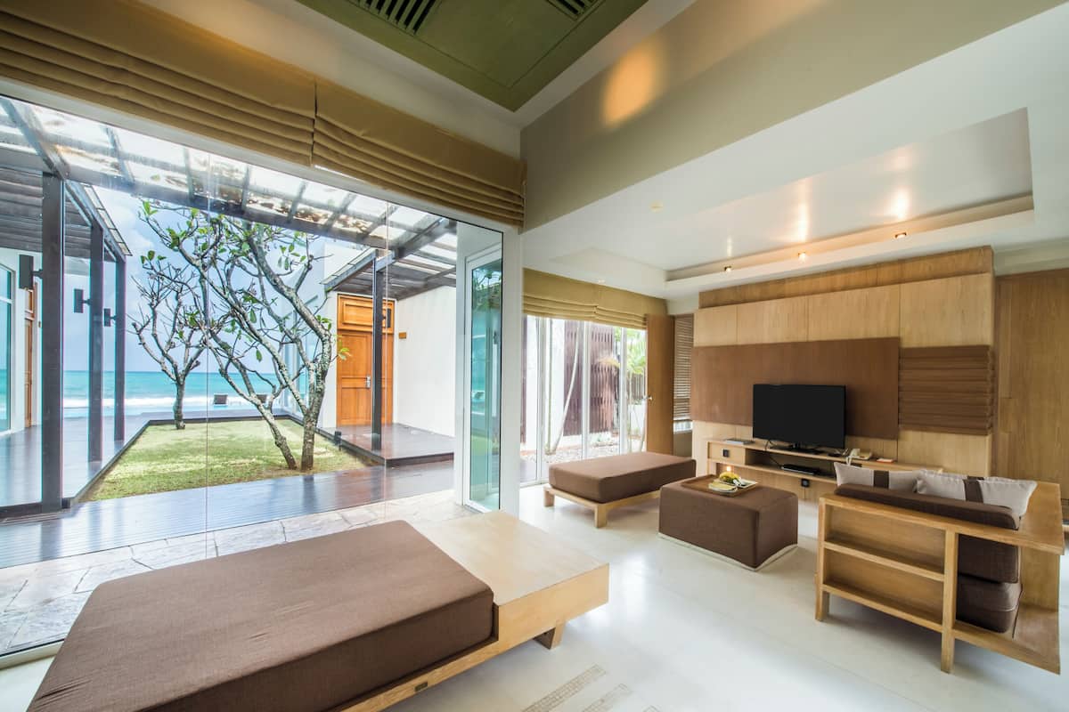 A modern living room with a large TV, wooden furniture, and a glass door leading to a balcony with a view of the ocean.