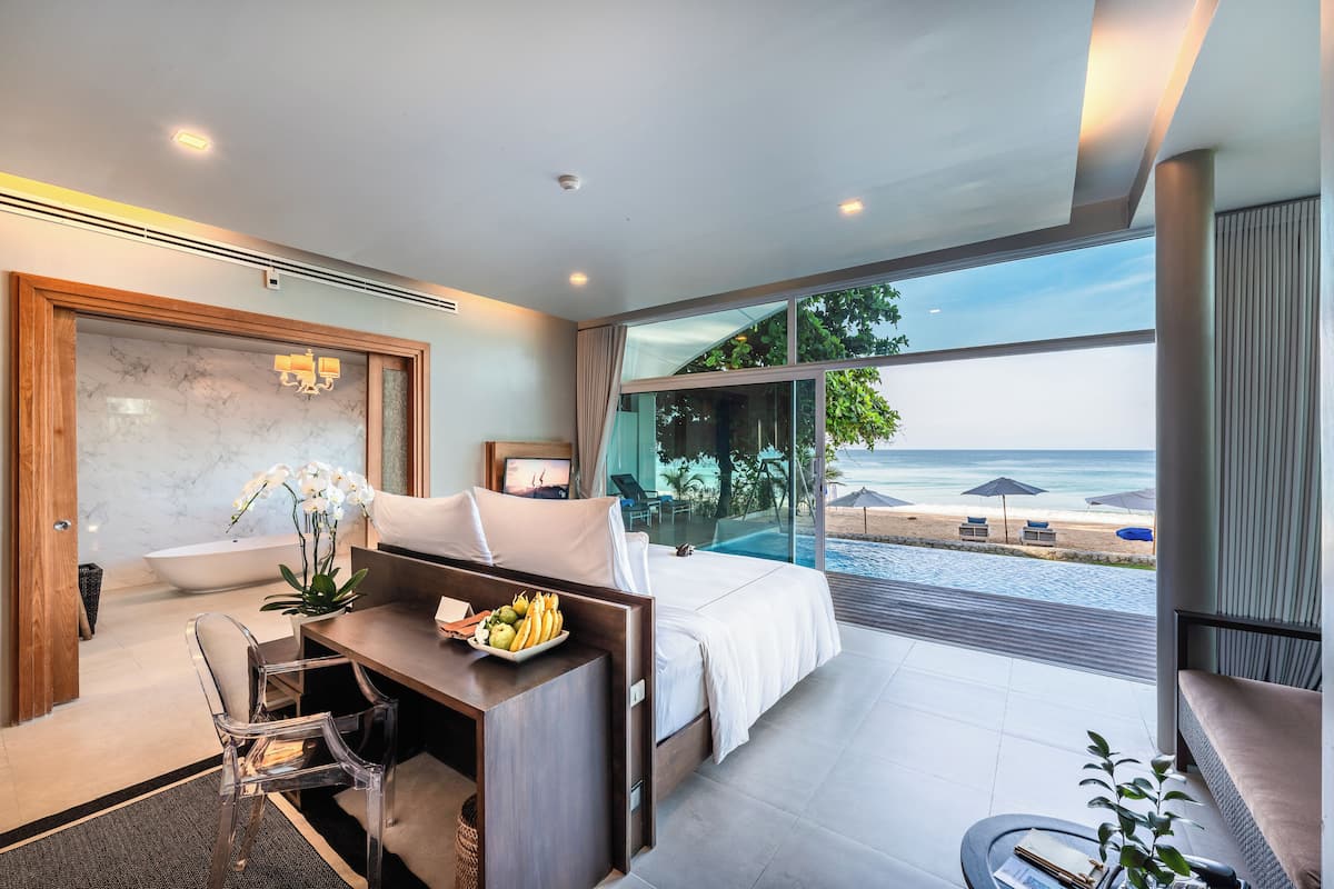 A modern hotel room with a large bed, a desk with a fruit bowl, a view of the beach, and a glass-enclosed balcony.