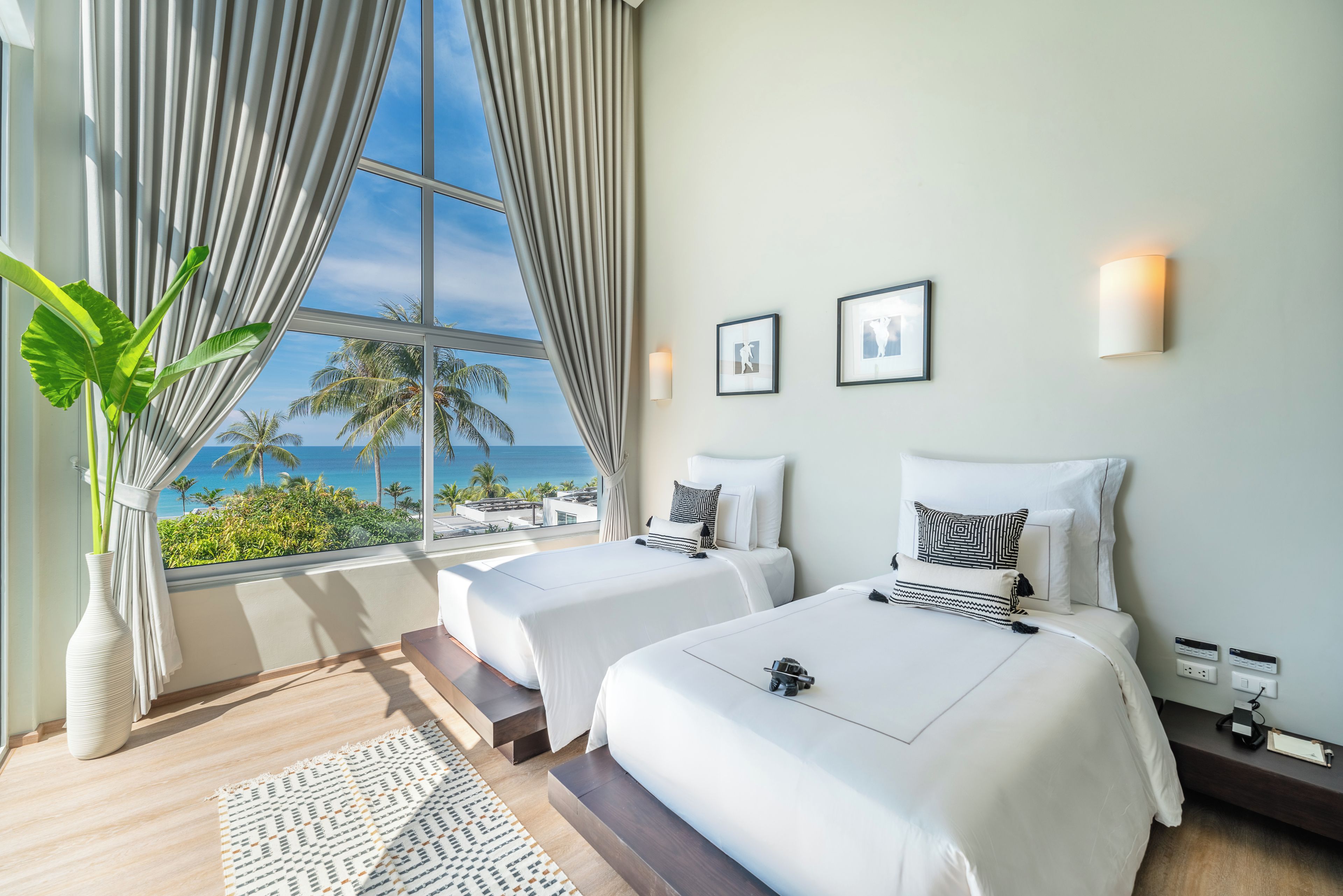 A hotel room with two beds, a large window with curtains, a plant, and a view of the ocean.