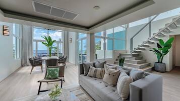 A spacious living room with a sofa, chairs, a dining table, and large windows offering a view of the ocean.