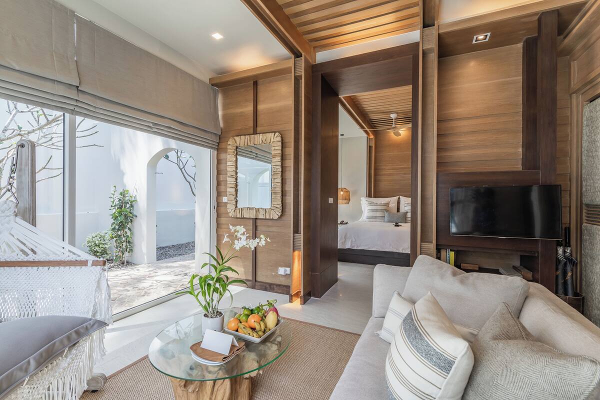 A modern hotel room with a large bed, a TV, a glass coffee table with fruit, a hammock, and a view of the outdoors.