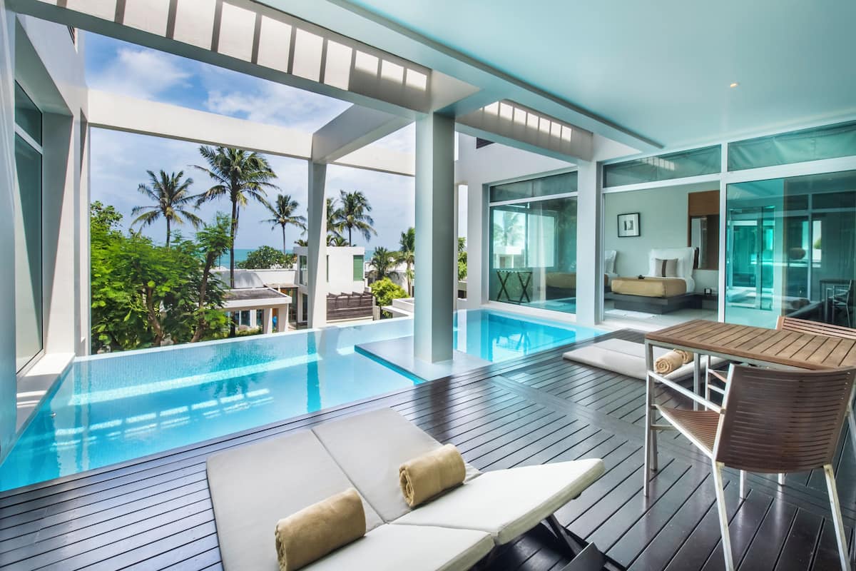 A modern infinity pool with a wooden deck, lounge chairs, and a glass-enclosed dining area with a view of palm trees and the ocean.