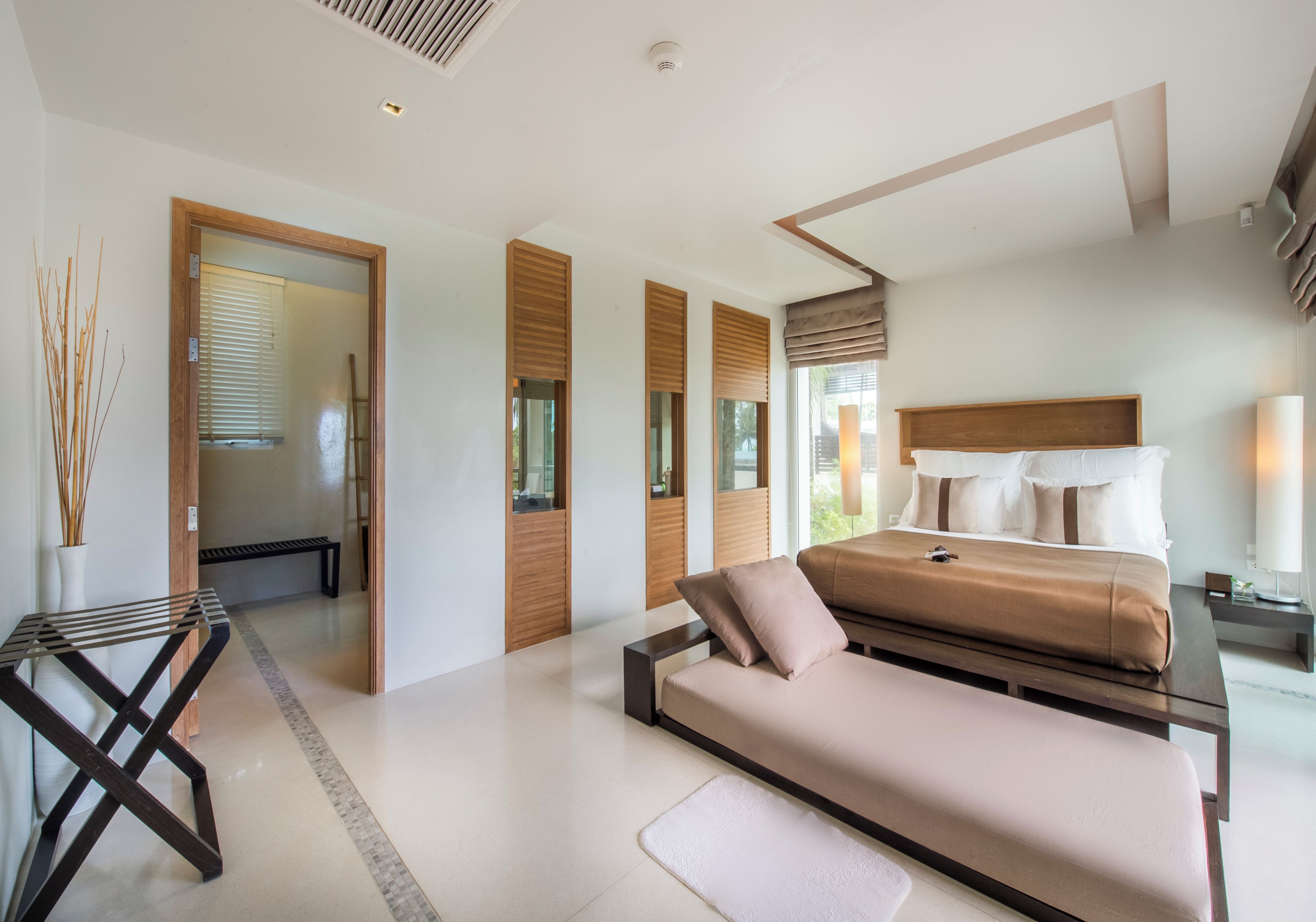 A modern bedroom with a bed, a bench, a small table, and a view of the outdoors.