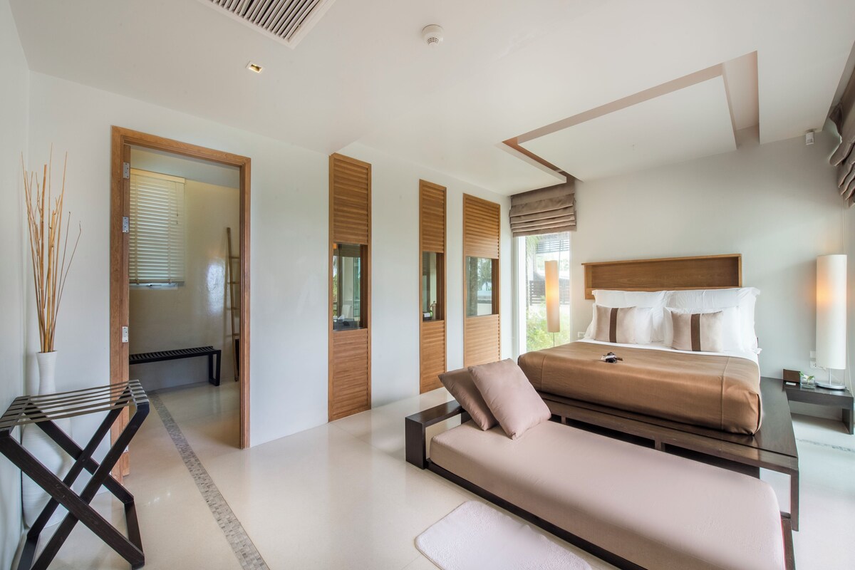 Quarto moderno com cama, um banco, uma mesinha e vista para o exterior.