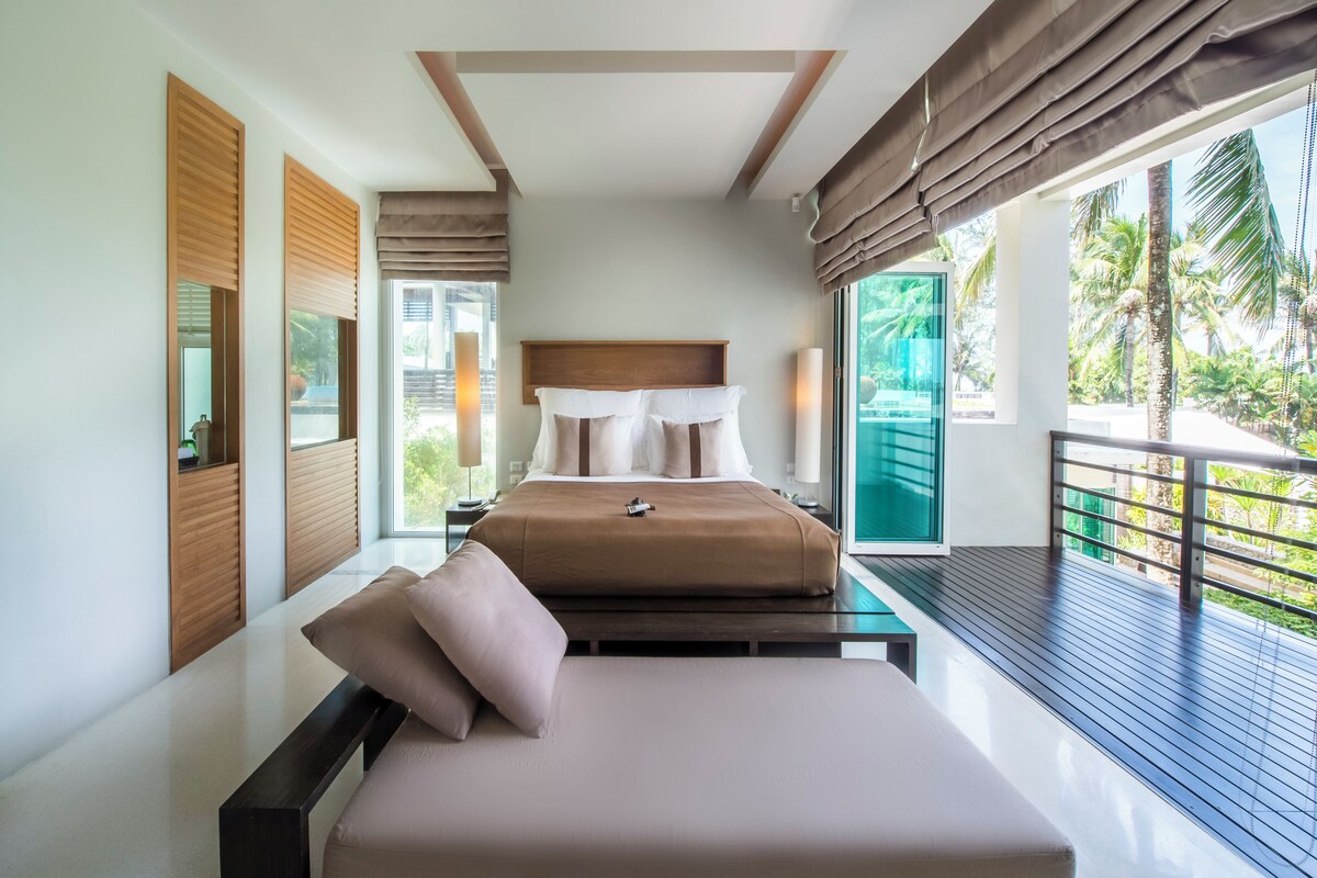 Quarto moderno com cama, mesas de cabeceira e varanda com vista para palmeiras.