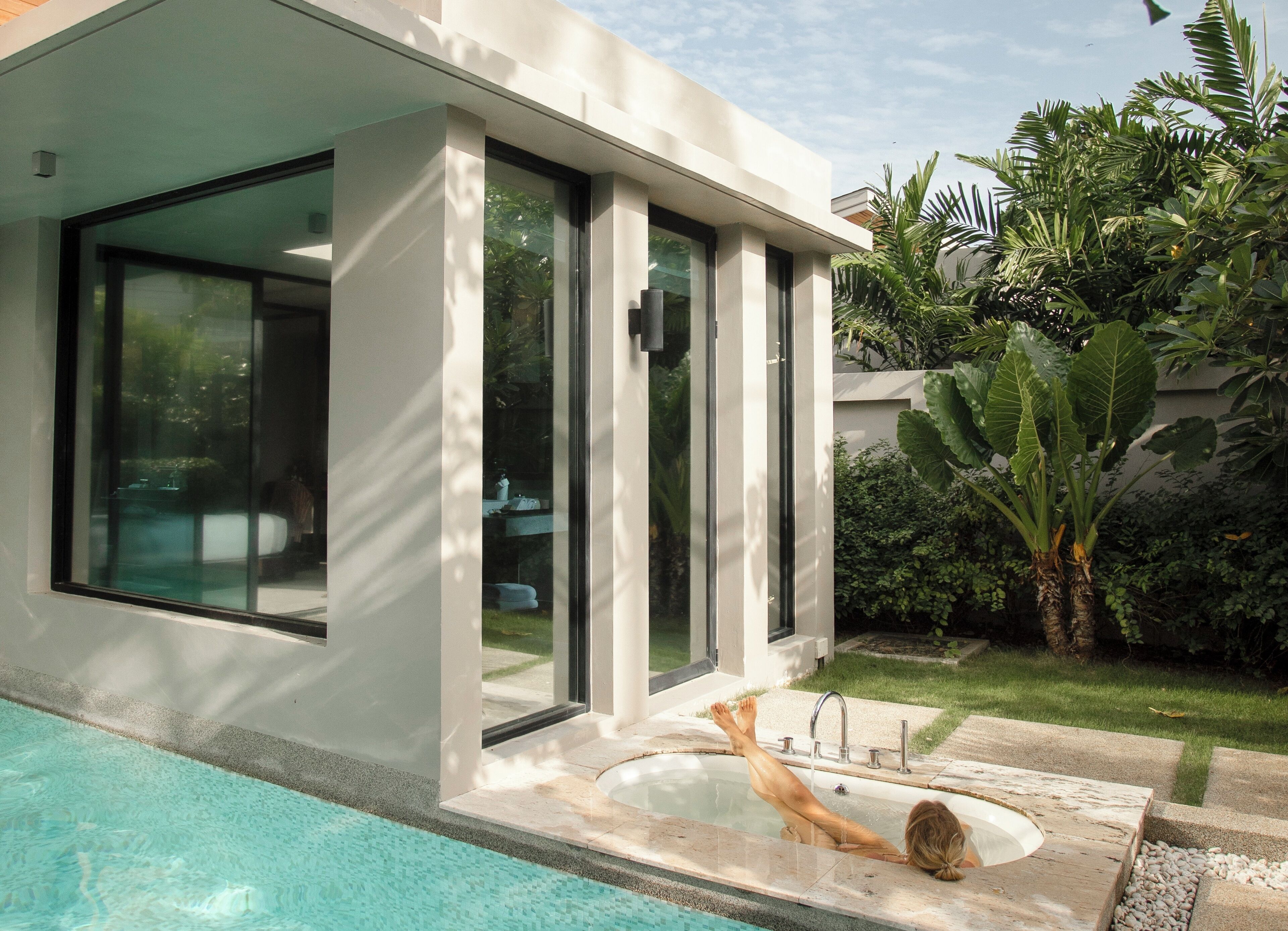 Ein modernes Haus mit Swimmingpool, großen Glasfenstern und einem Garten mit tropischen Pflanzen.