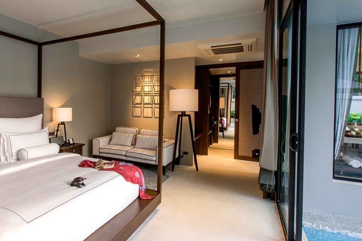 Quarto de hotel com cama grande, sofá, escrivaninha, luminária e vista para piscina.