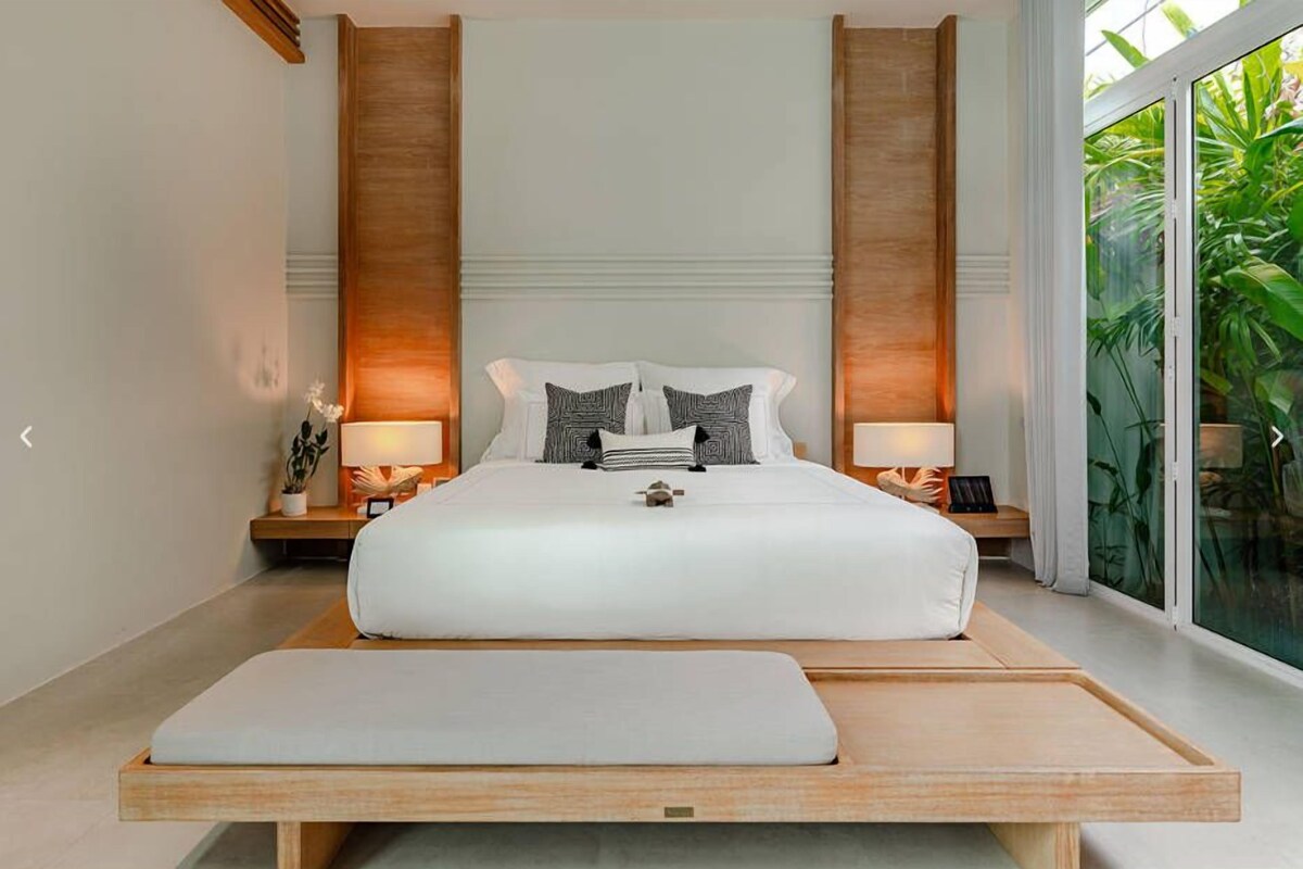 Quarto moderno com uma cama grande, mesas de cabeceira com luminárias e vista para o verde através de uma grande janela.
