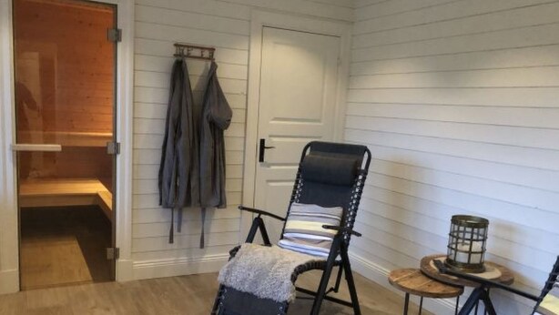 Une pièce avec un plancher en bois, une chaise noire avec un coussin, une petite table ronde avec une bougie et un porte-manteau avec des manteaux.