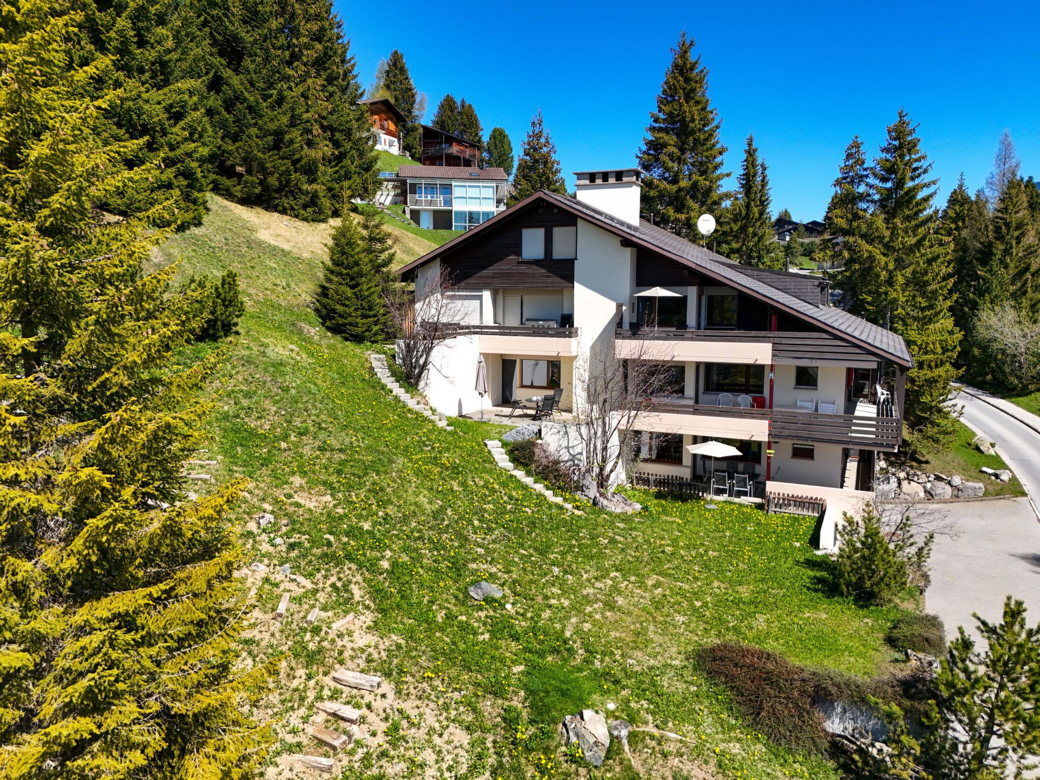 A modern two-story house with a balcony, surrounded by a grassy hillside and coniferous trees.
