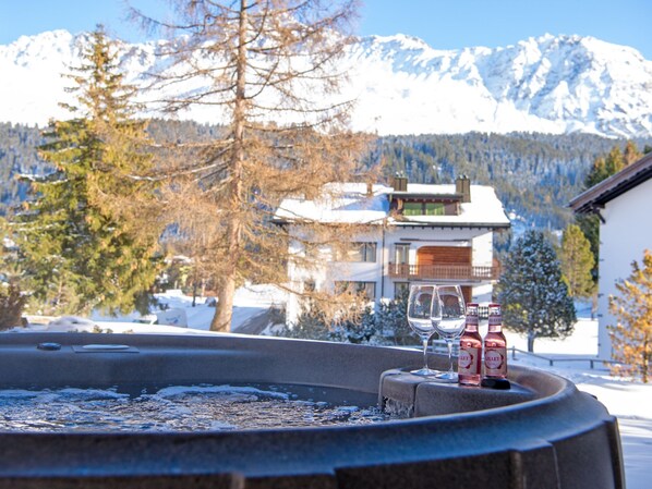 A snow-covered mountain landscape with a building featuring balconies and a pool area with two bottles of Lillet wine and two glasses.
