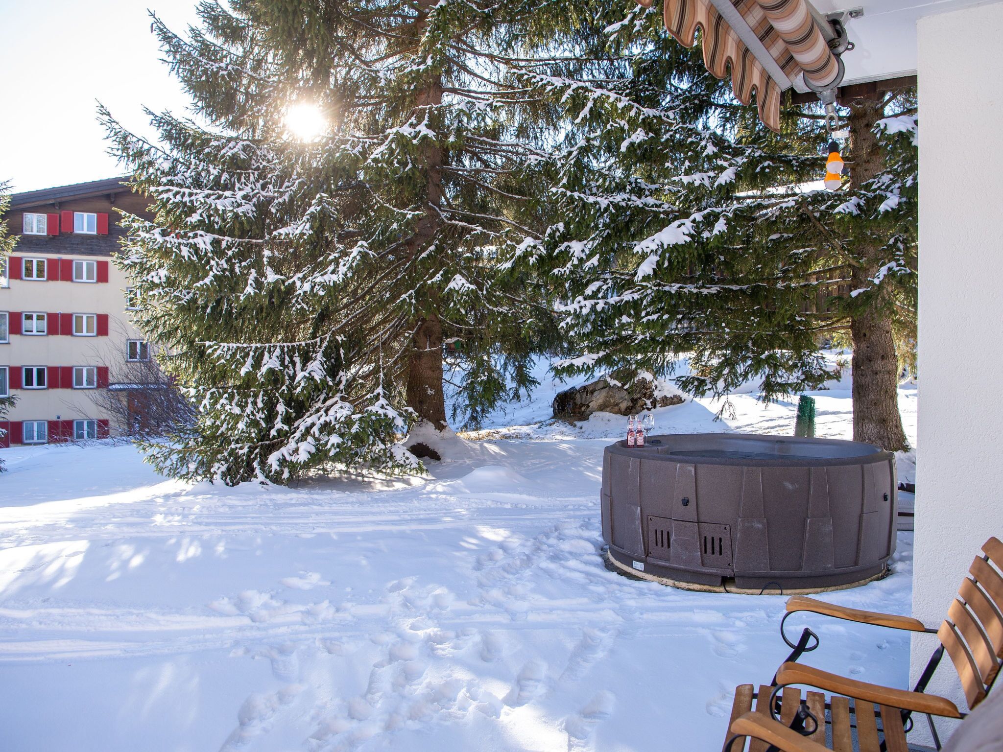 A snow-covered outdoor area with a hot tub, a wooden bench, and a building with red shutters.