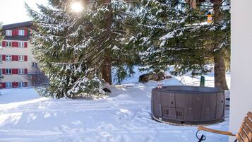A snow-covered outdoor area with a hot tub, a wooden bench, and a building with red shutters.