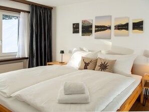 A hotel room with a large bed, white linens, a window with curtains, a nightstand with a lamp, and wall art depicting landscapes. - 1506 Casoja Valbella by Interhome (Valbella)