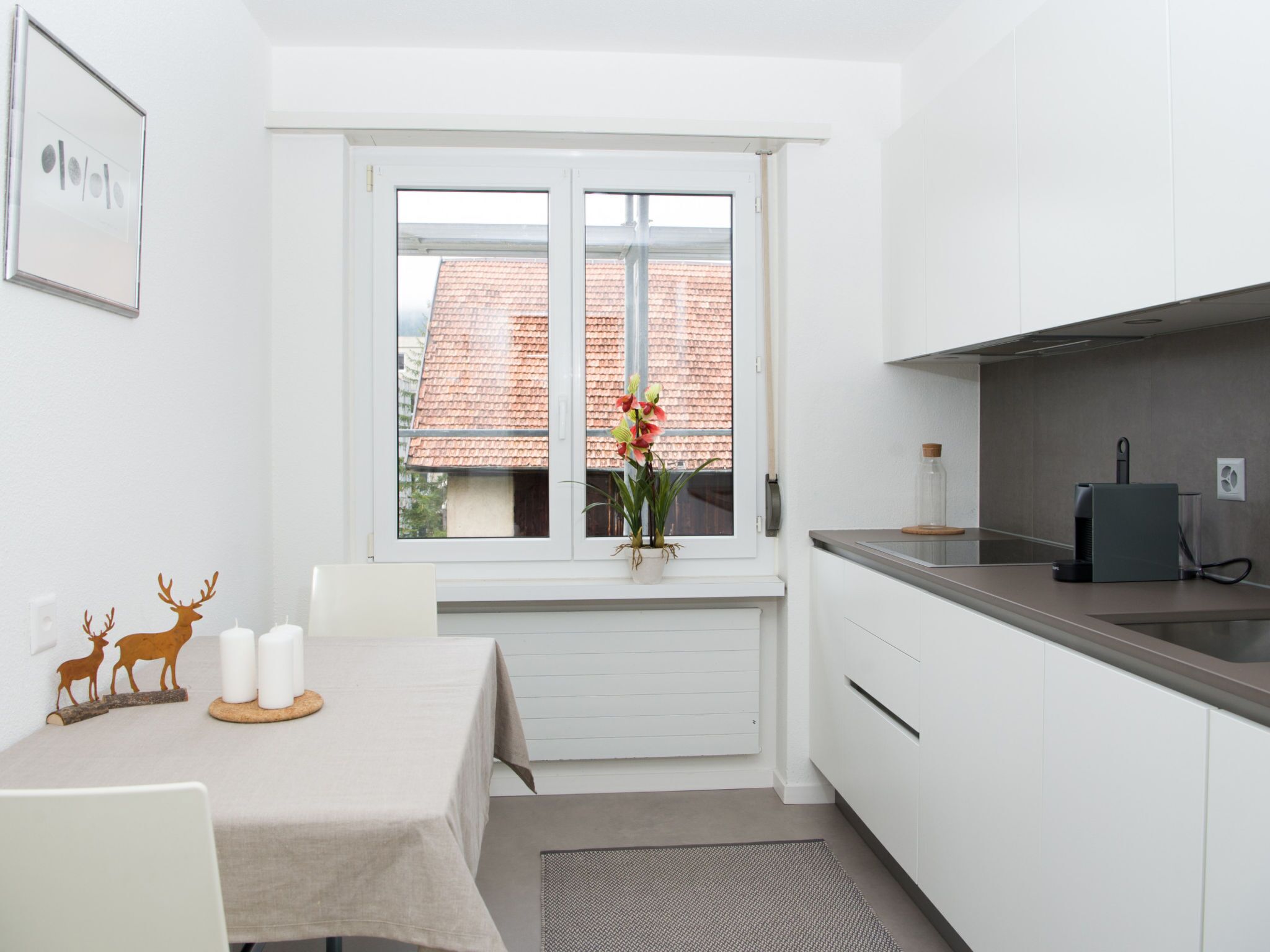 A small, modern kitchen with white cabinets, a stainless steel countertop, and a window with a view of a red-tiled roof.