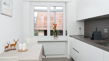 Eine kleine, moderne Küche mit weißen Schränken, einer Edelstahl-Arbeitsplatte und einem Fenster mit Blick auf ein rotes Ziegeldach.