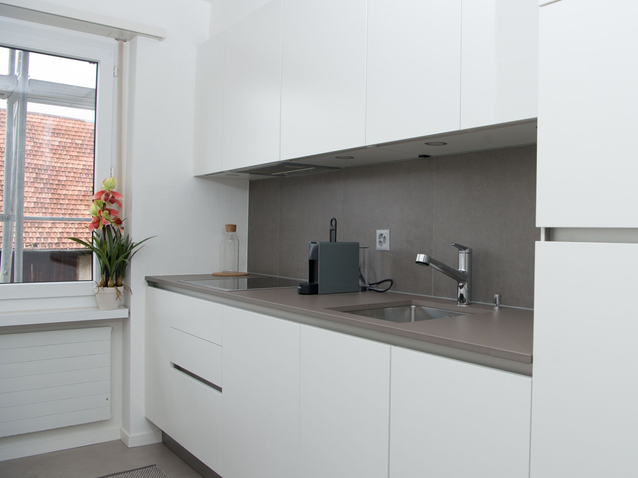 Eine moderne Küche mit weißen Schränken, einer grauen Arbeitsplatte, einem eingebauten Backofen und einem Fenster mit Blick auf ein Backsteinhaus.