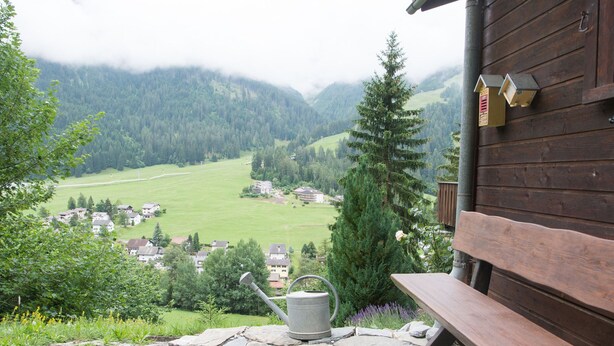 A stone patio with a metal watering can, a wooden bench, and a view of a valley with houses and mountains in the background. - 1301 Haus Joch by Interhome (Churwalden)