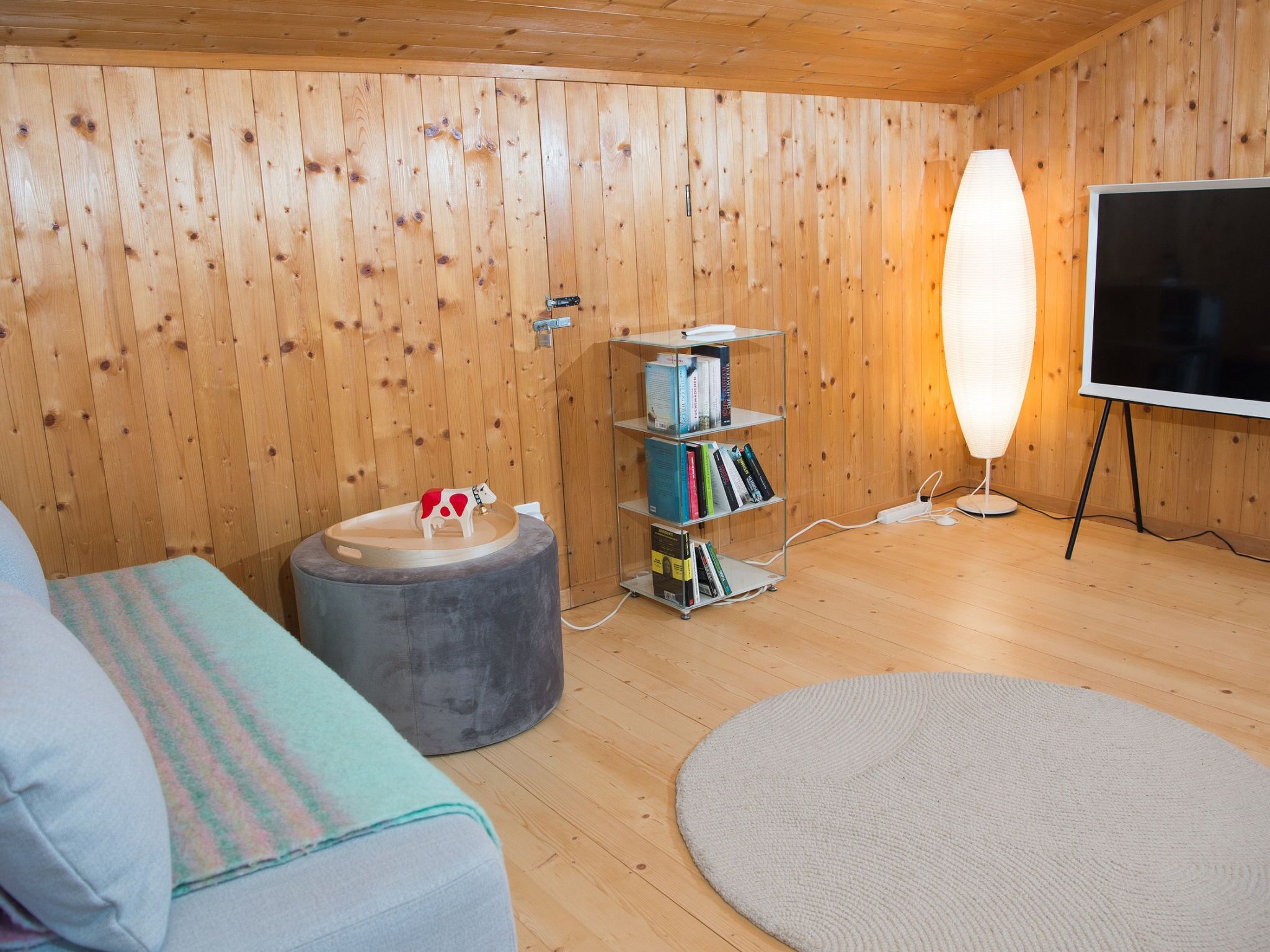Ein gemütliches Zimmer mit Holzwänden, einem runden Teppich, einer Couch, einem kleinen Hocker, einem Bücherregal und einem Fernseher auf einem Schrank.