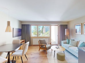 A modern living room with a dining area, a sofa, a coffee table, and a view of a mountainous landscape through large windows. - 1509 Chesa Barlangia Molinia by Interhome (Valbella)