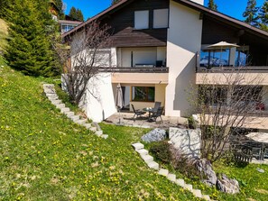 Una casa de dos pisos con balcón, un patio con mobiliario exterior y un jardín con flores amarillas.