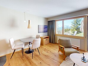 Ein modernes Zimmer mit Esstisch, zwei Stühlen, einer Couch, einem Fernseher und einem großen Fenster mit Blick auf die Berge.