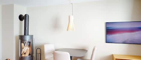 A modern living room with a wood-burning stove, a round dining table with chairs, a hanging light fixture, and a painting on the wall.