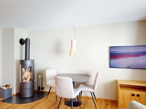 A modern living room with a wood-burning stove, a round dining table with chairs, a hanging light fixture, and a painting on the wall.