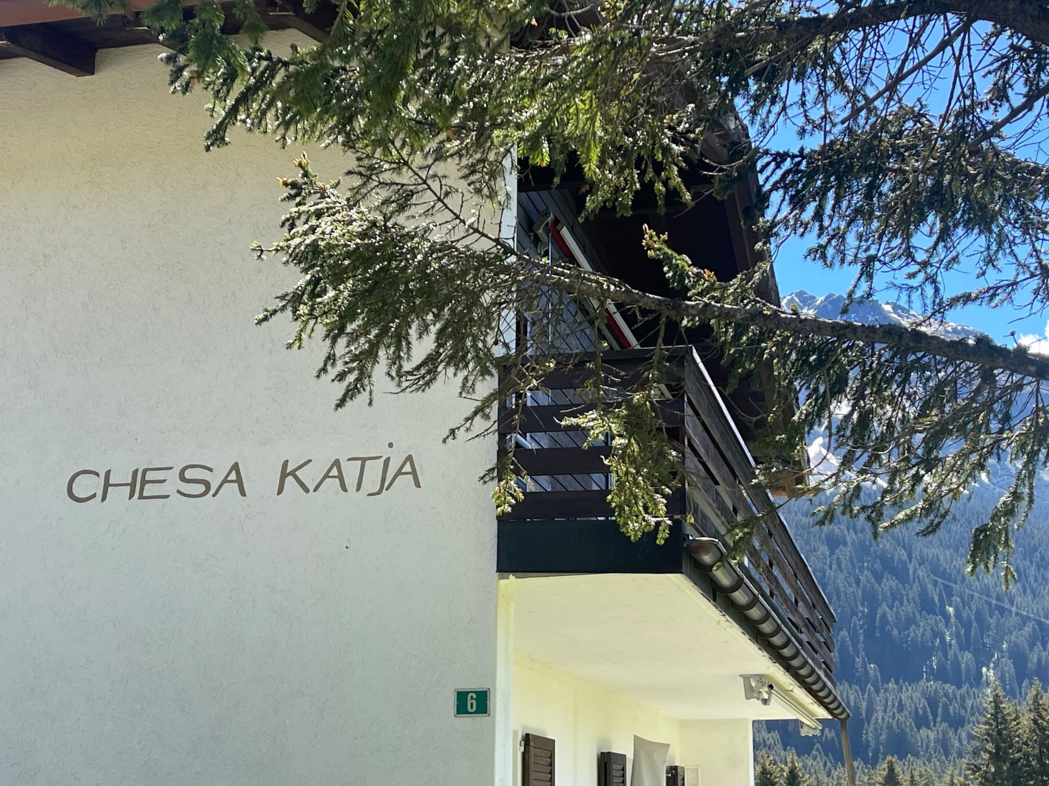 Un edificio con el nombre 'CHESA KATJA' en su fachada, una puerta marcada con el número '6', y una vista de montañas nevadas al fondo.